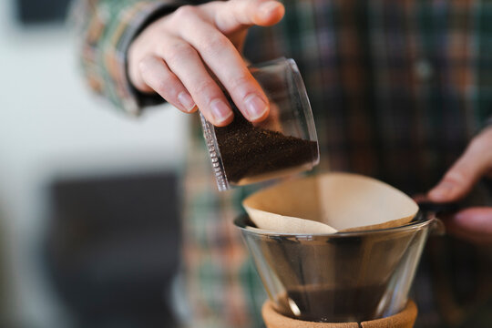 A person in a plaid shirt pours ground coffee from a small container into a paper filter inside a pour-over coffee maker. This is part of a morning brewing ritual.