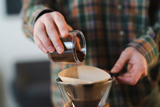 A person in a plaid shirt pours freshly ground coffee into a paper filter inside a glass pour-over coffee maker. This is part of the manual brewing process.