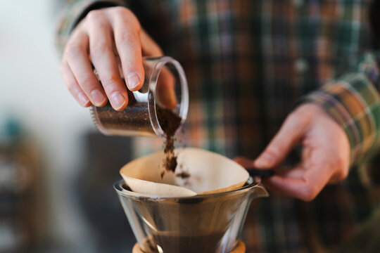 A person's hands pour freshly ground coffee into a paper filter inside a glass pour-over coffee maker. This is part of the morning brewing ritual.