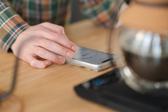 A person in a plaid shirt uses a smartphone, touching the screen. The phone displays a timer, used for brewing coffee with the device visible on the right.