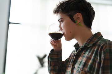A young adult with short, dark hair and a green ear gauge sips a dark beverage from a clear glass. The person wears a plaid shirt, enjoying a quiet moment indoors.