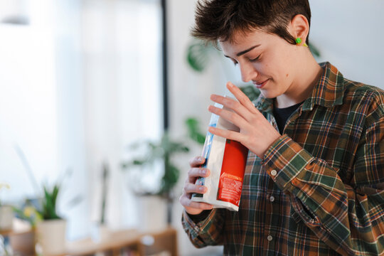 A young person in a plaid shirt holds a package, smelling its contents with a focused, content expression. They are enjoying the aroma of the product in a bright, modern indoor setting.