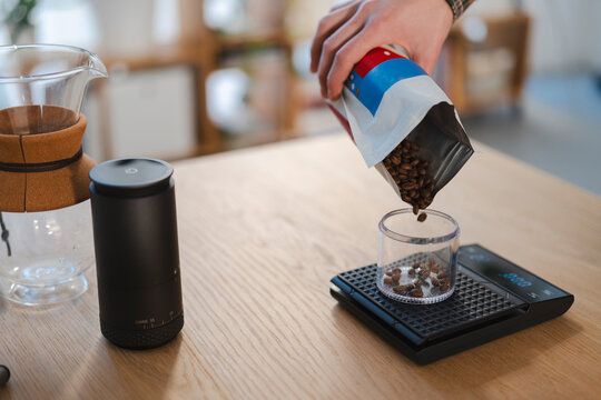 A person's hand carefully pours fresh roasted coffee beans from a bag into a clear container on a digital scale. This precise measurement is essential for a perfect pour-over coffee brewing ritual.