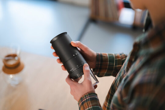 A person in a plaid shirt holds a black manual coffee grinder, carefully adjusting the grind size setting. They are preparing to brew fresh coffee at home.