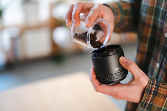 A person in a plaid shirt pours whole coffee beans from a clear cup into a black manual coffee grinder. They are preparing to grind fresh coffee for brewing.