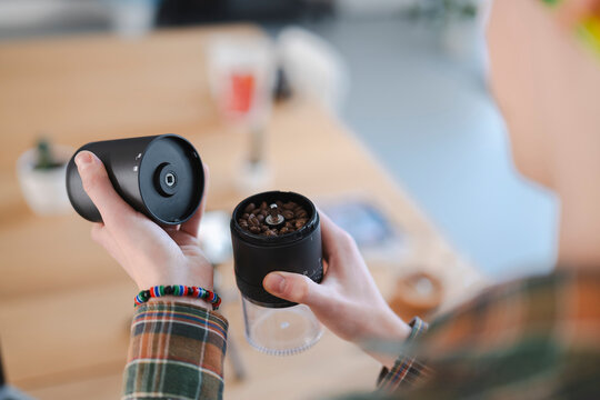 A person in a plaid shirt holds a manual coffee grinder filled with whole coffee beans, preparing to make fresh coffee. This captures the ritual of home brewing.
