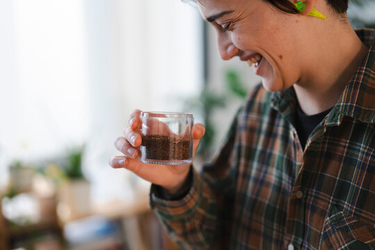 A happy young person in a plaid shirt smiles while holding a small clear container filled with dark coffee grounds. They are enjoying a moment of simple pleasure.