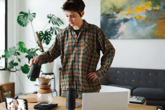 A young person in a plaid shirt carefully pours water into a pour-over coffee maker. They are filming the process with a smartphone and have a laptop nearby.
