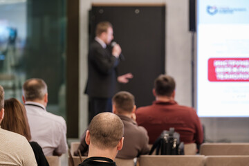 Audience seated facing speaker presenting at conference. Blurred presenter holds microphone while attention stays on stage. Modern venue with screen displays.