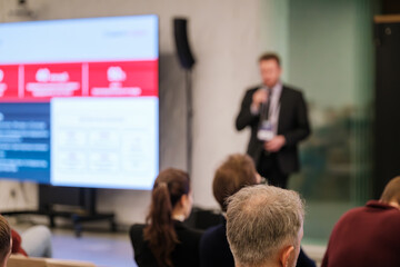 Presenter speaks at conference while audience watches large screen. Blurred background shows modern venue and event atmosphere. Focus on communication, business learning, professional gathering.