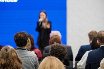 Speaker on stage with microphone addresses attendees. Blue backdrop creates vibrant conference setting. Audience sits attentive.