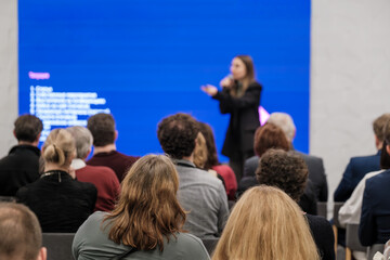 Audience listening to a speaker in a conference setting with a bright blue screen. Attentive crowd in rows creating a professional event mood.
