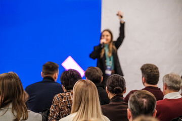 Conference scene with female speaker on stage microphone in hand audience listening intently blue backdrop creative lighting energy engagement.