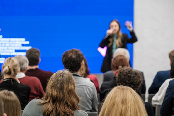Presenter speaks to attentive crowd during corporate conference. Bright blue backdrop highlights stage presence while audience listens, notes, engages. 