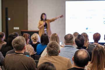 Presenter speaks to attentive audience during conference session with large screen slides. Energetic tone, informative content, professional environment, collaboration and learning emphasized.