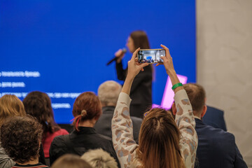 Audience watches speaker on stage. Different people in foreground record with smartphone. Blue screen backdrop. Presenting event atmosphere energetic and engaging. Focus on phone screen.