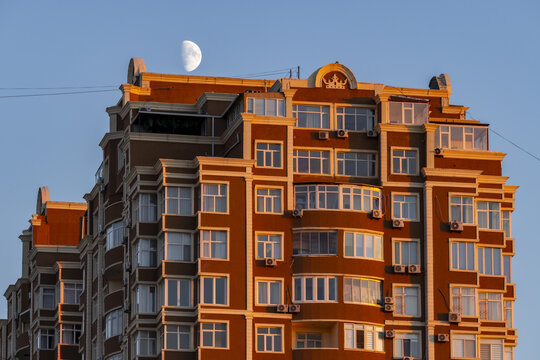 View of a tall, red-brick building with many windows stands bathed in the warm glow of sunset beneath a half moon in the clear sky, Aktau, Mangystau Region, Kazakhstan.