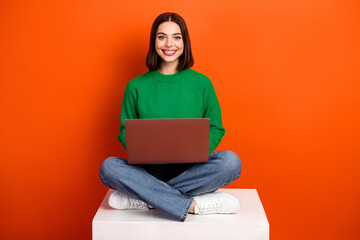 Young chic student sits cross legged on a white platform with a laptop smiling against an orange background