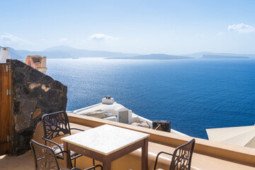 View of a serene outdoor dining area with wrought iron chairs and a table overlooking the vast, deep blue sea under a bright sky, Oia, Thira, Greece.