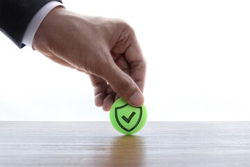 A hand places a green circle with a drawn shield and checkmark icon on a wooden table. Security, safety, insurance, protection, verification concept.