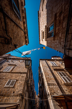 View of historic stone buildings rise towards a vibrant blue sky in a narrow street, dotted with flags and lamps, creating a unique urban canyon, Dubrovnik, Dubrovnik-Neretva County, Croatia.