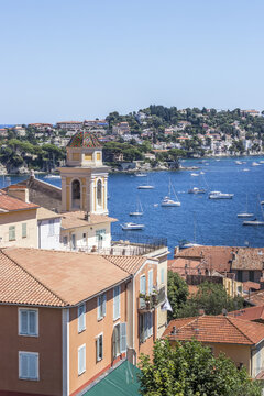 View of terracotta rooftops cascading towards the azure sea dotted with white sailboats, a charming bell tower piercing the skyline, Villefranche-sur-Mer, Provence-Alpes-Cote d'Azur, France.