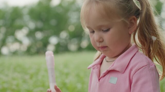 caucasian girl blows soap bubble with wand in sunlit field, concentrated breath and gentle grip shimmering bubbles, soft summer grass, reflective expression, tiny bow in ponytail, intimate candid