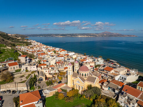 Aerial view of sun-kissed buildings cascade towards the glittering sea, dominated by a church with a pink dome, creating a tranquil coastal vista, Kalyves, Chania, Greece.