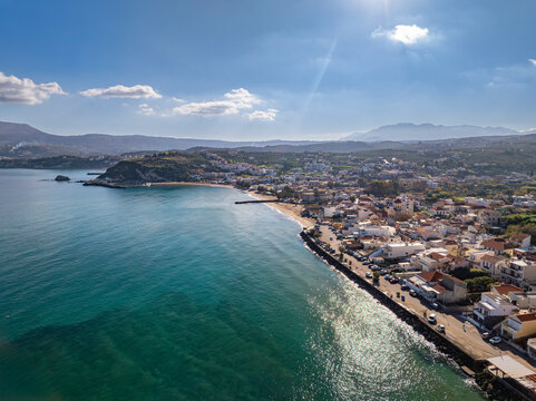 Aerial view of turquoise waters meet sandy shores and white-washed buildings under a clear blue sky, creating a stunning contrast of colors in Kalyves, Chania, Greece.