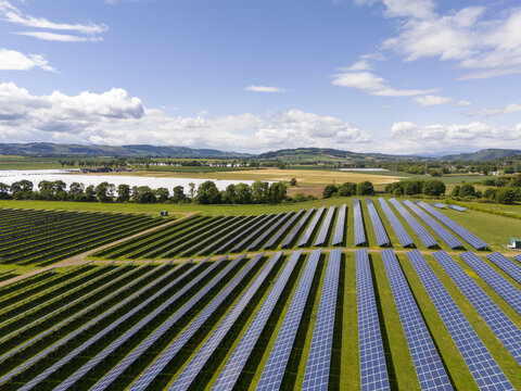 Aerial view of rows of solar panels stretch across the landscape, reflecting the sky's azure hues, a contrast to the green fields beyond, Perth, Scotland, United Kingdom.