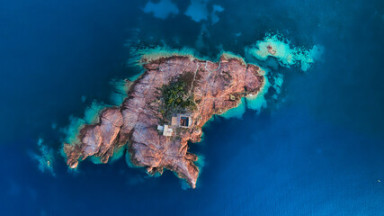 Aerial view of a rugged, rust-colored island standing in stark contrast to the deep blue sea, a small structure nestled amongst the rocks, St RapahaÃ«l, France.