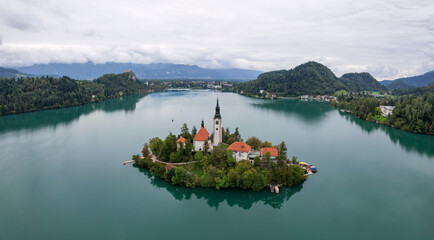 Aerial view of a quaint island church with a dark spire rising amidst the turquoise lake, framed by lush green forests, Bled, Slovenia.