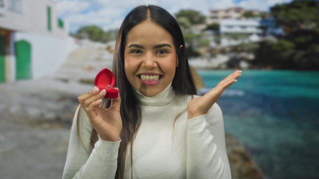 Woman holding engagement ring box, smiling with hand to mouth, standing on a scenic beach, showcasing excitement for an outdoor proposal.