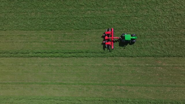 Aerial view shows green tractor pulling red mower in rural farmland during daytime harvest. Title in description. Operator is visible. Top down composition and linear rows.