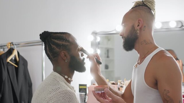 Makeup artist applying foundation to gay man face in dressing room with mirror lights in studio