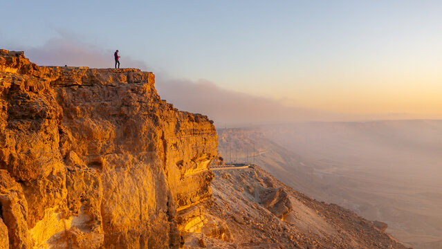 Aerial view of a lone figure stands silhouetted against the horizon atop the Mitzpe Ramon crater rim, bathed in the warm glow of sunrise, Mitzpe Ramon, South District, Israel.