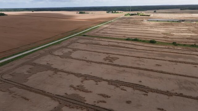 Aerial pan of harvested wheat strips, service road, mast, farm. Tractor tracks and stacked swaths show methodical harvest rows and subtle parallax across terrain.
