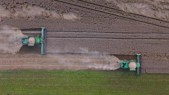 Aerial view of two combine harvesters cutting ripened cereal fields. Title appears here. Machines move side by side, carve parallel lines, and raise dust plumes.