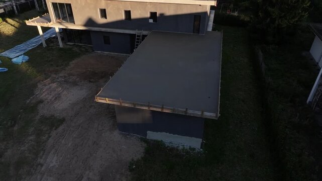 Aerial drone approach shows flat roof residence under construction, concrete slab, wooden formwork, cantilevered slab, ladder, debris, solar panels, conifers, late afternoon light.