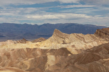 Fototapeta premium A panoramic vista unfolds at Zabriskie Point, where deeply eroded badlands stretch across the foreground in waves of pale gold, tan, and ash gray.