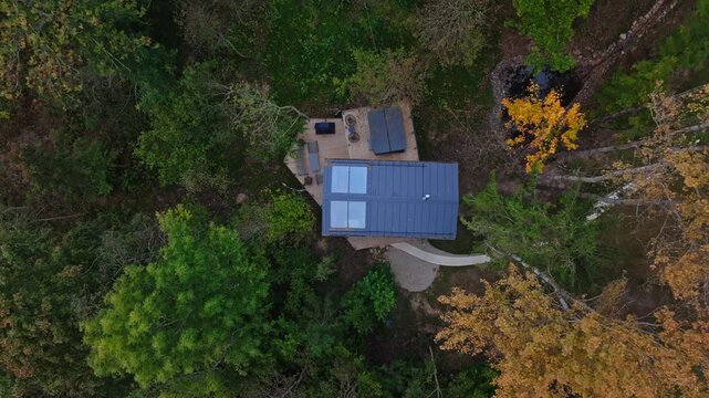 Aerial view shows modern tiny house with metal roof and two skylights on a triangular wooden deck beside a curved stone path and small pond in mixed forest, slow pan and rotation.