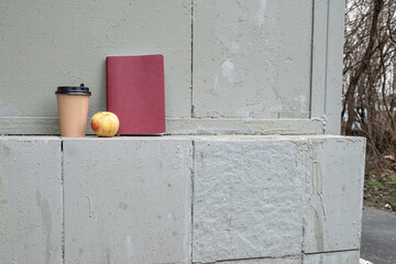 Paper coffee cup, notebook, and apple placed on rough concrete ledge outdoors, symbolizing quick study break, remote work pause, and urban education or coworking lifestyle visuals