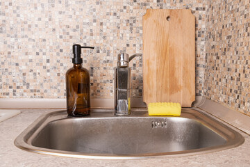 Kitchen sink with refillable soap dispenser, sponge, and wooden cutting board against mosaic backsplash, illustrating everyday dishwashing routine