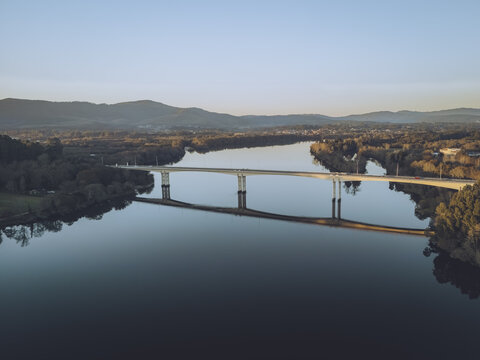 Aerial view of a bridge stretching across a tranquil river, reflecting the clear sky, framed by verdant trees and distant mountains, Vila Nova de Cerveira, Viana do Castelo District, Portugal.
