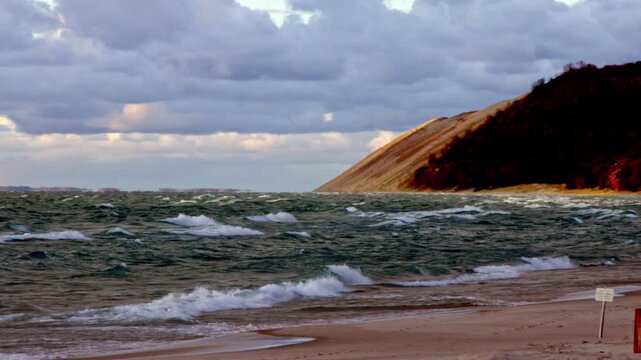 Lake Michigan&rsquo;s powerful waves crash under a cloudy autumn sky, with Sleeping Bear Dunes rising in the background for a dramatic fall shoreline view wide shot pan left to right.