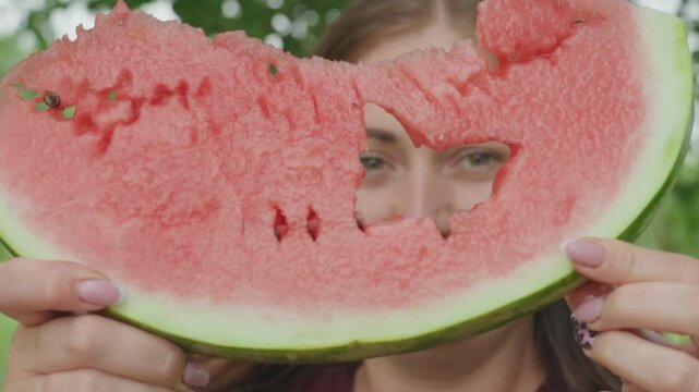 white woman holds watermelon slice outdoors, peeking through heartshaped bite, playful smile, green backyard, summer picnic vibe, juicy red flesh with seeds, thick green rind, closeup portrait