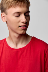 Young man in a red shirt looks down with a calm, contemplative expression in a clean studio setting, showcasing casual style and thoughtful mood suitable for fashion, lifestyle, or editorial stock