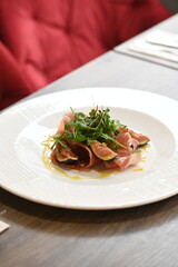close-up view of served food, sliced Parma ham, fresh arugula and figs with dressing, light plate on restaurant table, elegant gourmet appetizer,