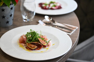 close-up view of served food, sliced Parma ham, fresh arugula and figs with dressing, light plate on restaurant table, elegant gourmet appetizer,