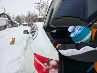 Man cleaning snow from car trunk in winter village street, with a dog sitting nearby. Cold season and travel preparation.